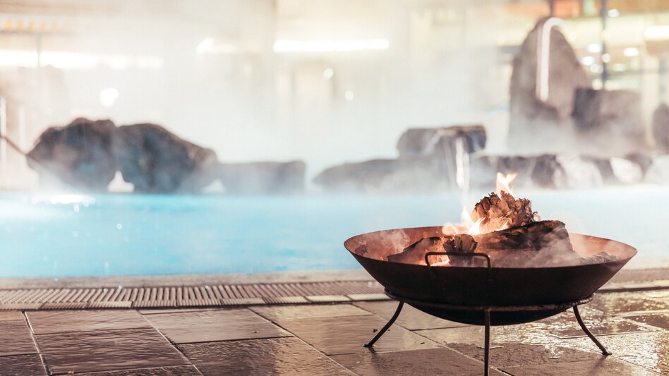 Knisterndes Feuer in Feuerschale am Rand eines dampfenden Außenpools im Thermenresort Loipersdorf, stimmungsvolle Wellnessatmosphäre im Winter mit Wasser, Felsen und warmem Licht.