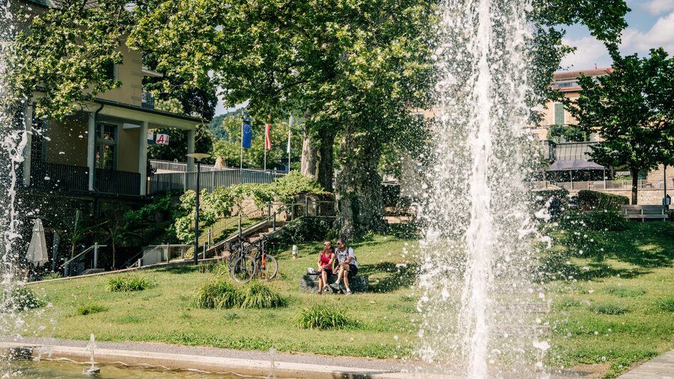 Wasserspiel im grünen Kurpark von Bad Gleichenberg im Thermen- & Vulkanland Steiermark mit hohen Wasserfontänen und schattenspendenden Bäumen.