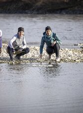 Drei Frauen beim Winterspaziergang an der Mur im Thermen- & Vulkanland Steiermark, kniend am Flussufer mit Kieseln, ruhige Naturstimmung an einem klaren Tag.