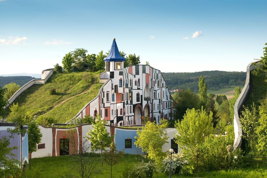 Verspieltes, farbenfrohes Gebäude der Therme Rogner Bad Blumau im unverkennbaren Stil von Friedensreich Hundertwasser, eingebettet in die grüne Hügellandschaft der Oststeiermark. | © Rogner Bad Blumau, Hundertwasser Architekturprojekt