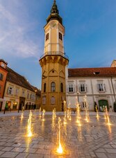 Beleuchteter Hauptplatz mit dem historischen Rathausturm von Bad Radkersburg und den leuchtenden Wasserspielen am Abend – stimmungsvolle Altstadt im Thermen- & Vulkanland Steiermark.