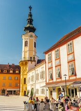 Altstadt von Bad Radkersburg mit historischem Kirchturm, farbenfrohen Fassaden und Gästen in einem Straßencafé bei sonnigem Wetter im Thermen- & Vulkanland Steiermark.