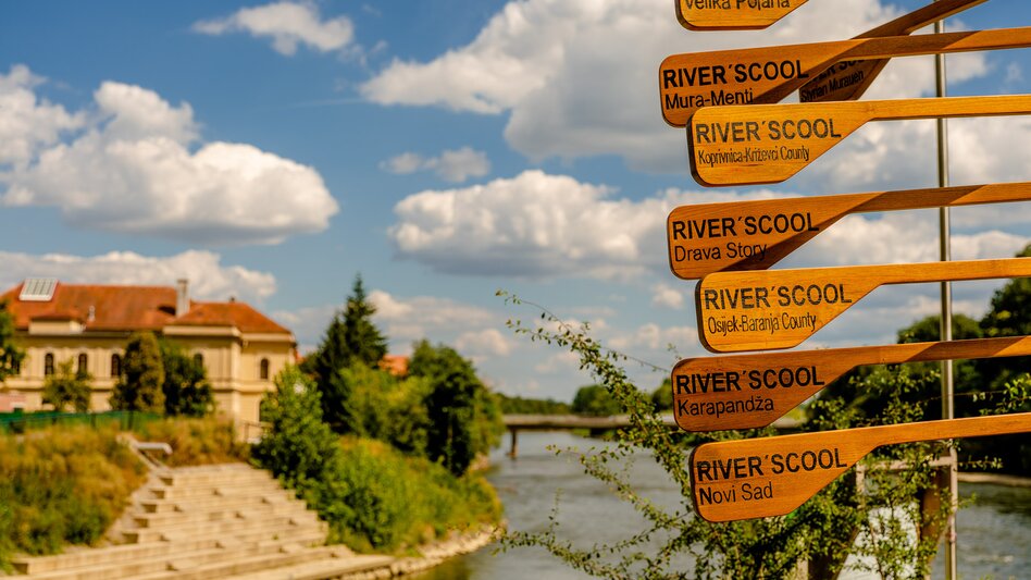 River School an der Mur in Bad Radkersburg mit internationalem Wegweiser am Flussufer und Blick auf die Mur in der historischen Kurstadt in der Südoststeiermark.