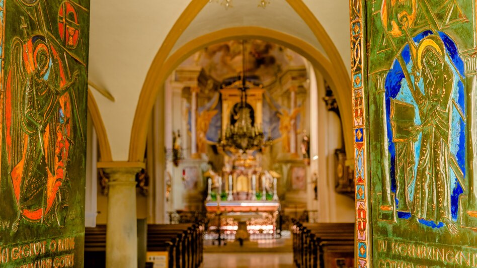 Kircheninnenraum in Bad Radkersburg mit kunstvoll gestalteten Eingangstüren, gotischem Spitzbogen, Kirchenbänken und reich verziertem Altar.
