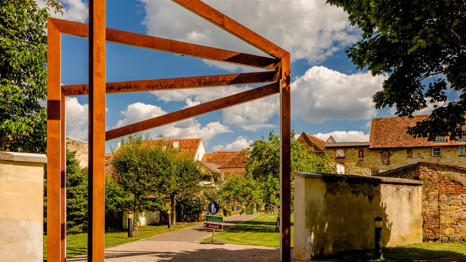 Markante Stahlkonstruktion in der historischen Altstadt von Bad Radkersburg, umgeben von alten Mauern, grünen Bäumen und traditioneller Architektur.