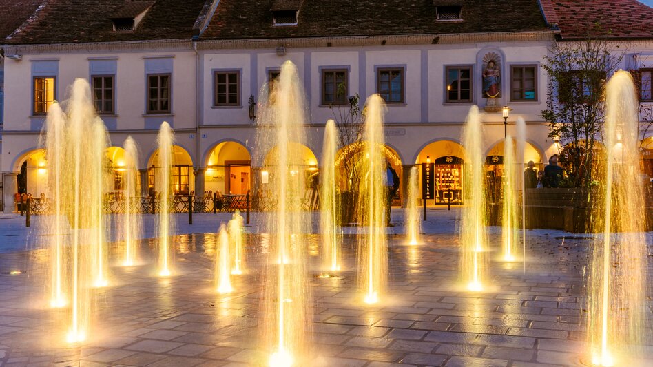 Beleuchtetes Wasserspiel in der Altstadt von Bad Radkersburg mit zahlreichen Fontänen vor historischen Gebäuden und Arkaden in der Abenddämmerung.
