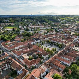 Luftaufnahme der historischen Altstadt von Fürstenfeld im Thermen- & Vulkanland Steiermark mit geschlossenem Häuserensemble, rotdachigen Bürgerhäusern, Hauptplatz und umliegender grüner Hügellandschaft an einem sonnigen Tag.