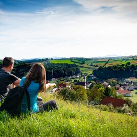 Two women and a man sit on the green, lush meadow and look out over Unterlamm in Thermen- & Vulkanland Styria