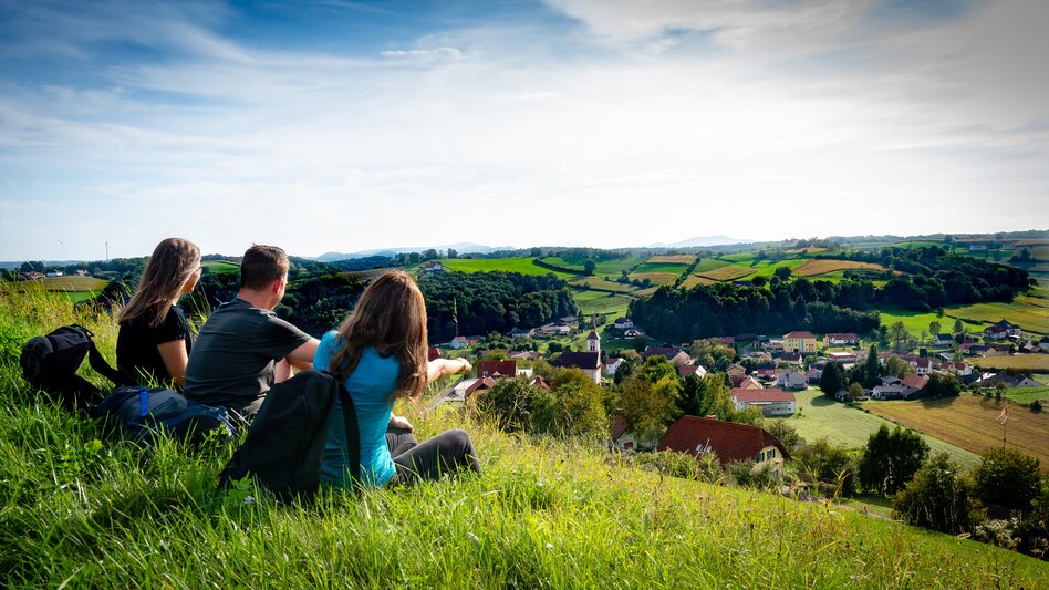 Zwei Frauen und ein Mann sitzen auf der grünen, saftigen Wiese und blicken auf Unterlamm im Thermen- & Vulkanland Steiermark