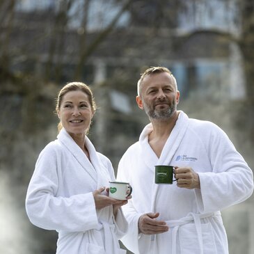 Paar im Bademantel und mit Kaffeetassen vor der Therme