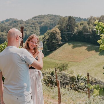 Weinverkostung auf einer Terrasse in einem Weinberg mit Blick auf eine bewaldete Hügellandschaft. Zwei Gäste stoßen mit Weißwein an und genießen den Sommerurlaub im Thermen- & Vulkanland.