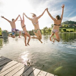 Freundesgruppe in Badekleidung springt von Steg in Naturbadesee in Riegersburg