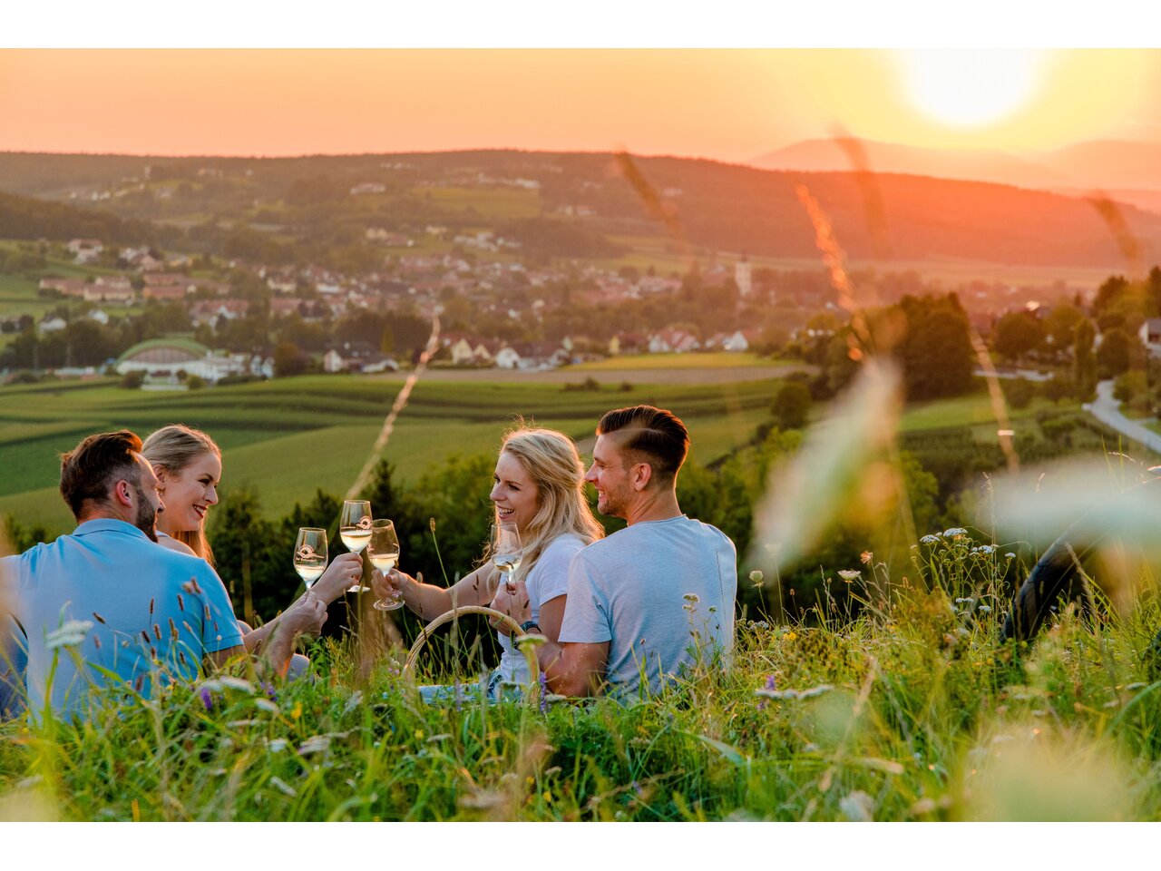 Vier Freunde sitzen auf der Wiese und stoßen mit Weingläsern bei Sonnenuntergang an