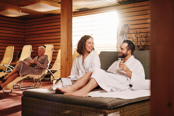 Relaxační místnost moderního wellness a spa centra s hosty v županech odpočívajícími na pohodlných lehátkách. Dřevěná architektura vytváří klidnou atmosféru pro relaxaci a regeneraci.