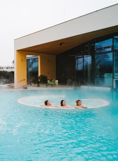 Außenbereich einer Thermen- und Wellnessanlage mit Thermalbecken, Wassermassageelement und modernem Spa-Gebäude. Drei Badegäste entspannen im warmen Wasser eines ganzjährig nutzbaren Outdoor-Pools.
