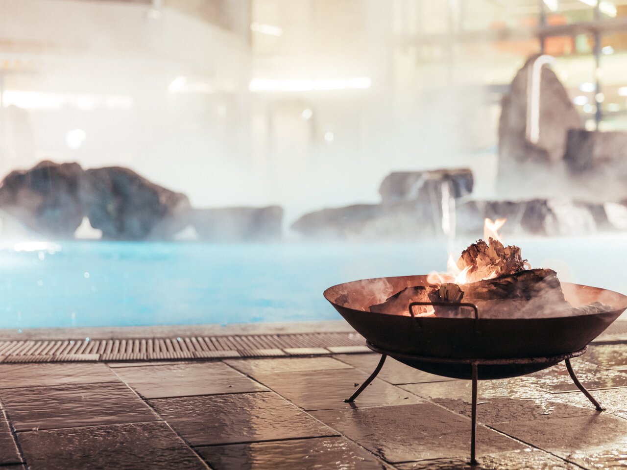 Z teplého termálního bazénu Thermenresort Loipersdorf stoupá pára. V popředí hoří ohniště na mokré kamenné dlažbě, zatímco v pozadí stéká voda po skalách a vytváří klidnou wellness atmosféru.