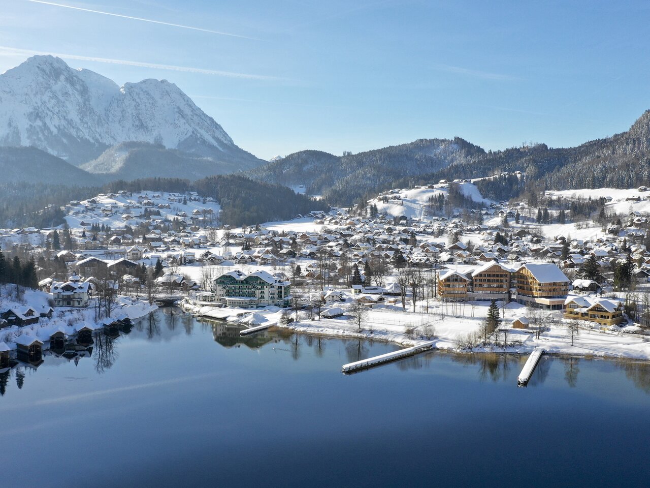Winterliche Luftaufnahme von Altaussee: Der tiefblaue See liegt ruhig zwischen schneebedeckten Bergen, umgeben von verschneiten Wäldern und dem kleinen Ort mit seinen Häusern am Ufer. | © Tom Lamm