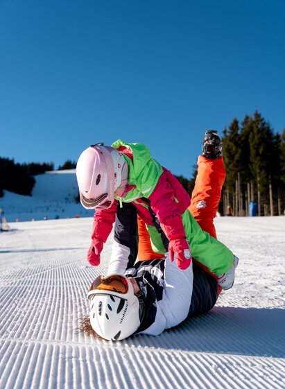 Ein Kind steht auf einem Erwachsenen im Schnee, beide lachen und tragen Skiausrüstung.