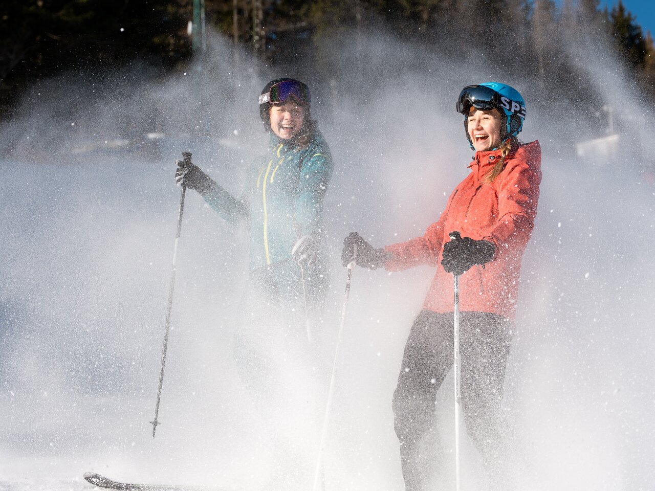Zwei Frauen stehen auf Skiern im Schnee, umgeben von Neuschnee und Bäumen.