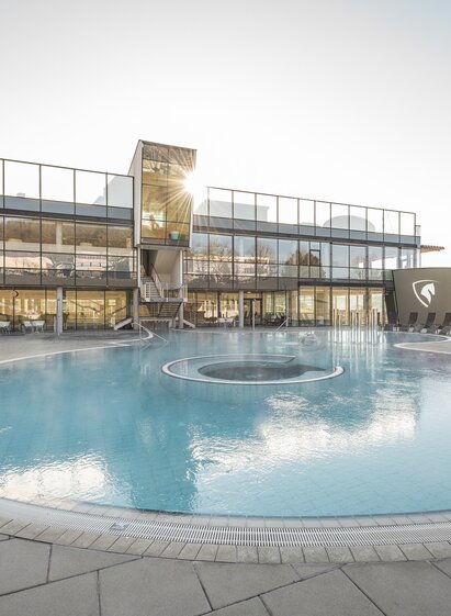 Moderne Therme mit großer Außenpoollandschaft, umgeben von Liegestühlen und Glasfassaden, in einer natürlichen Umgebung mit sanften Hügeln im Hintergrund. Das warme Wasser dampft leicht in der kühlen Luft.
