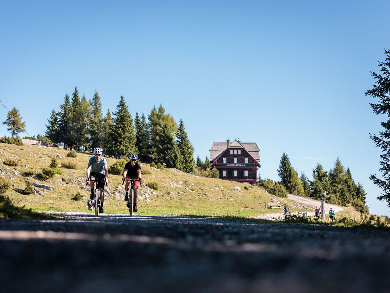 Zwei Radfahrer auf einer Bergstraße am Schöckl, im Hintergrund eine Hütte und Wanderer in alpiner Landschaft  EN