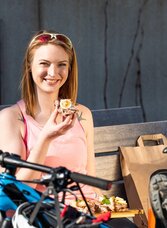 Radfahrerin genießt ein regionales Schmankerl auf einer Bank während der Schmankerl Tour