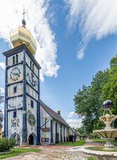 Die von Friedensreich Hundertwasser gestaltete St. Barbara Kirche in Bärnbach mit einem markanten, bunten Turm und einem Brunnen im Vordergrund.