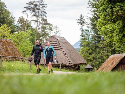 Two cyclists in cycling gear walking through the Austrian Open-Air Museum Stübing by the Mur cycle path, with traditional farmhouses and greenery in the background
