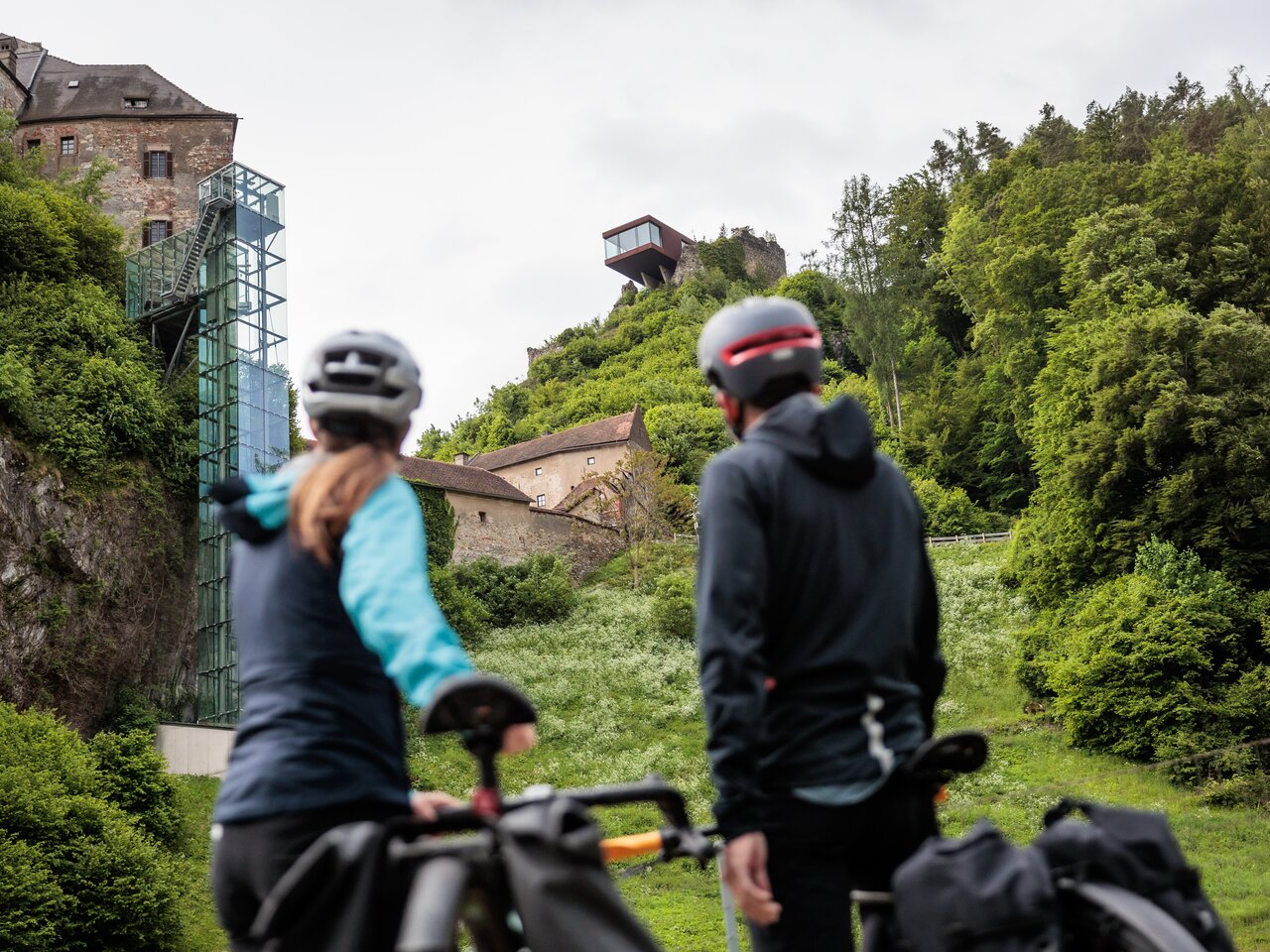 Two cyclists standing next to their bikes looking at Rabenstein Castle with a glass lift, surrounded by greenery