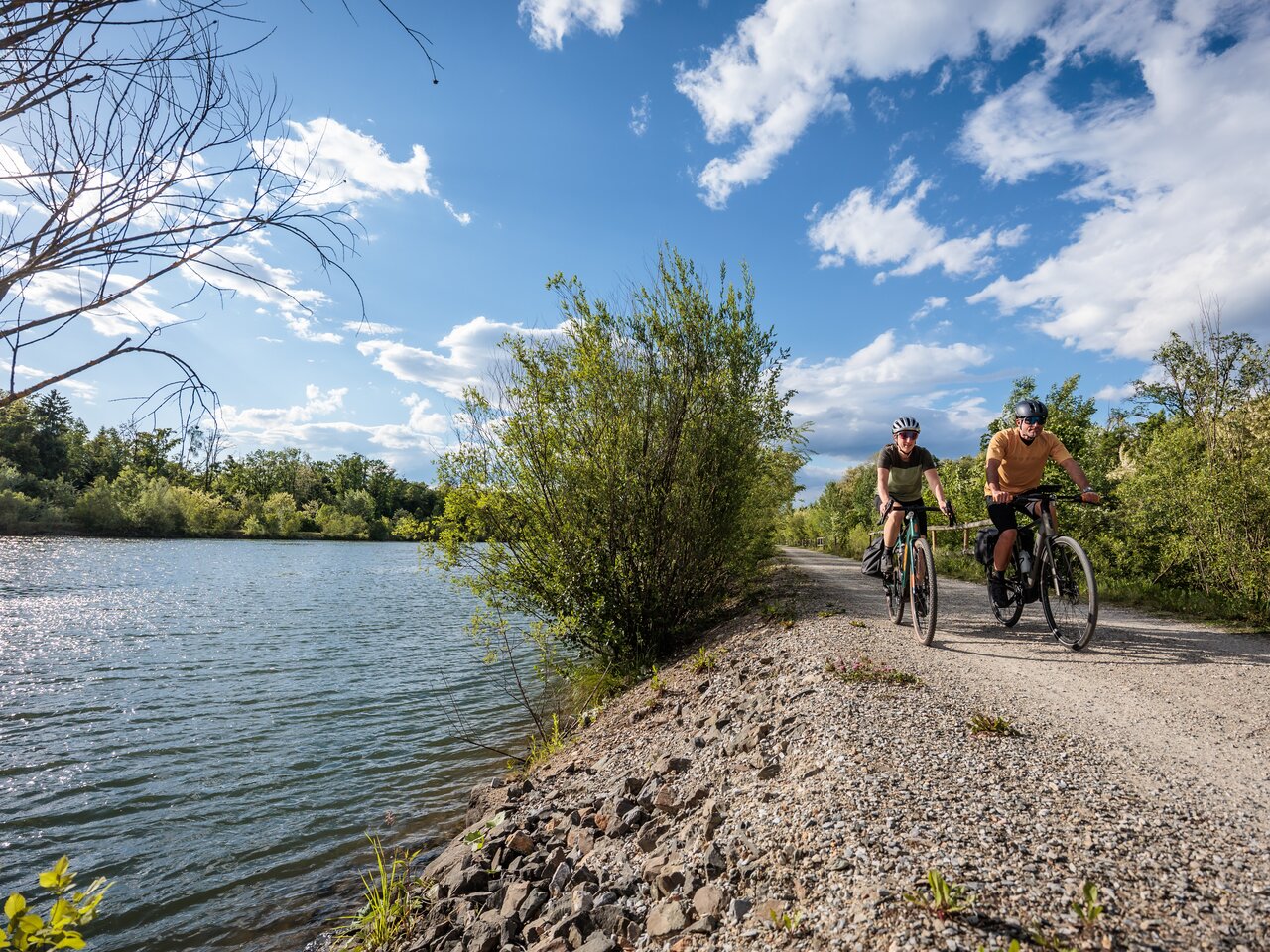 Zwei Radfahrer fahren auf einem Schotterweg entlang der Mur durch die grünen Murauen bei Kalsdorf bei Graz