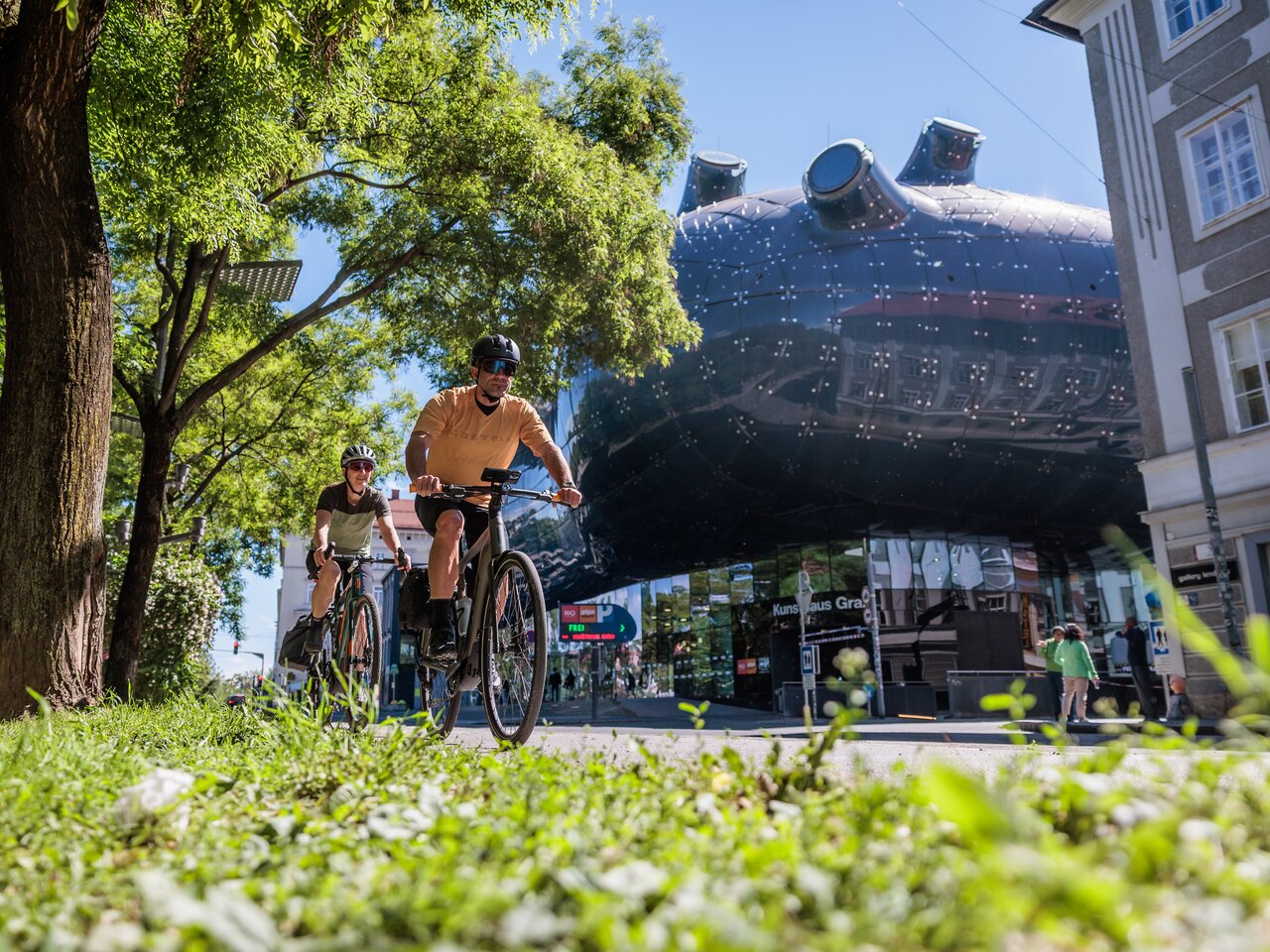 Two cyclists on a bike path in Graz with Kunsthaus Graz in the background in summer