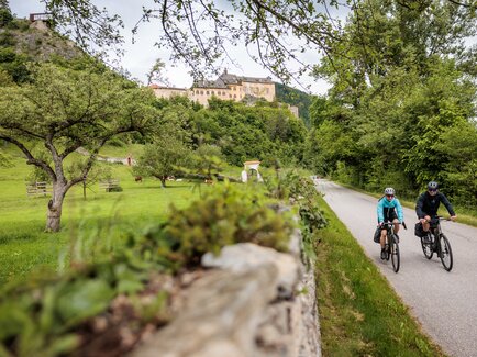 Two cyclists on the Mur cycle path with Rabenstein Castle in the background, surrounded by lush greenery