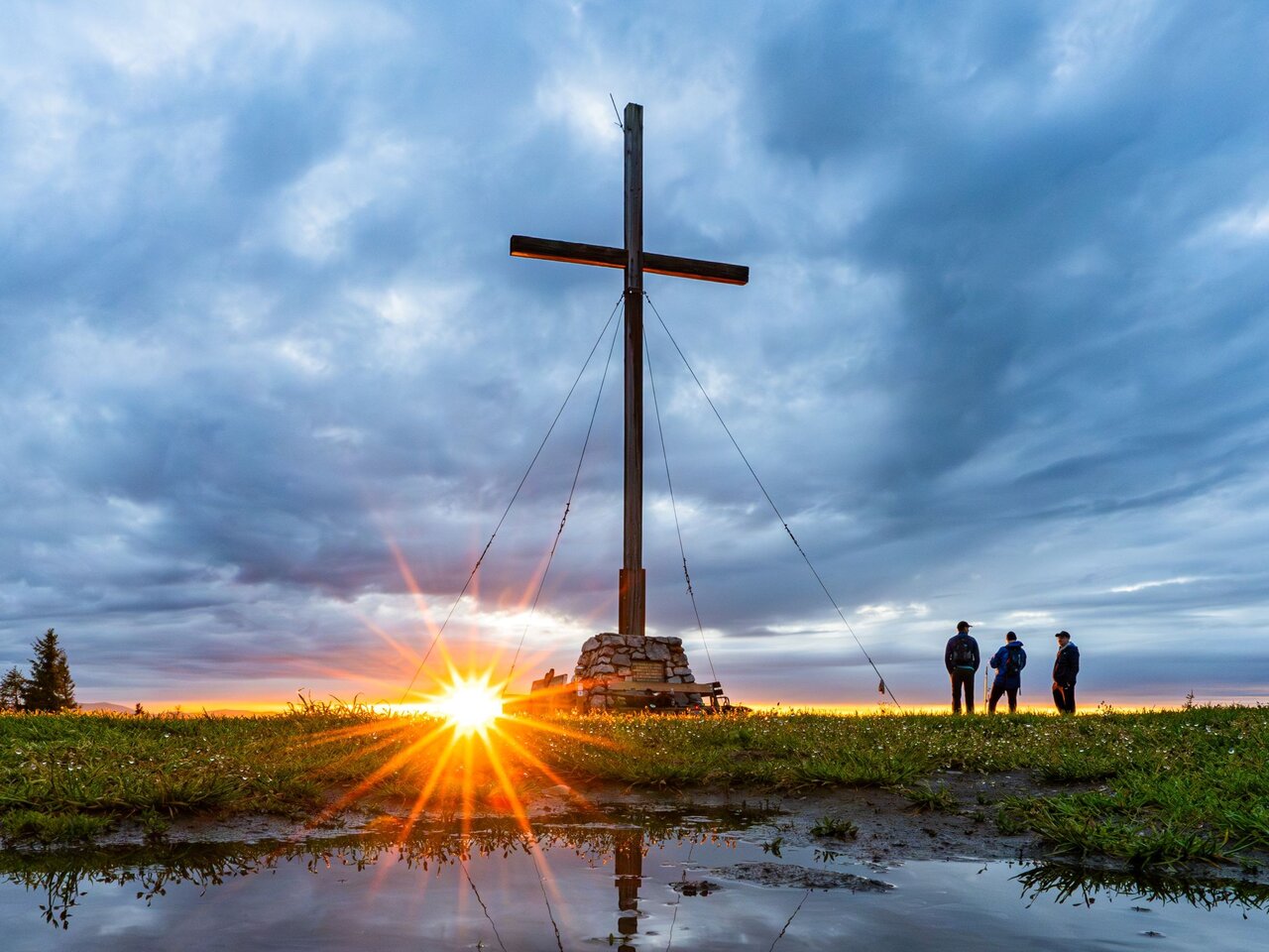 Sonnenaufgang am Gipfel des Schöckl mit Gipfelkreuz, Wandernden und weitem Blick über die Region Graz.