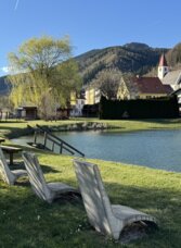 Pastner Teich in Übelbach mit Sitzplätzen am Ufer und Blick auf den Ort und die umliegenden Berge.
