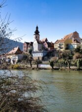 Blick über die Mur auf die historische Altstadt von Frohnleiten mit bunten Häusern und Kirchturm.