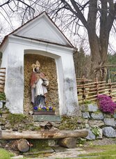 Ulrichsbrunn in Semriach mit Heiligenfigur, Steinmauer und traditionellem Holzgeländer.