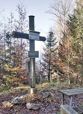 Gipfelkreuz am Hochtrötsch bei Semriach mit Sitzbank, Laub und Waldlandschaft.