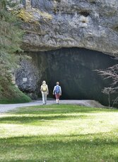 Zwei Wanderinnen vor dem Eingang zur Lurgrotte Semriach, umgeben von Felsen, Wald und Wiesen.