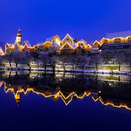 Die schneebedeckte Altstadt von Frohnleiten erstrahlt in festlicher Beleuchtung entlang der Mur, deren ruhige Wasserfläche die winterliche Szenerie in der Dämmerung spiegelbildlich reflektiert.