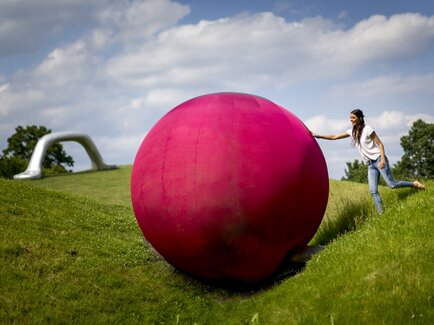 A woman pushing a large red sphere sculpture in the Sculpture Park Graz on a green meadow