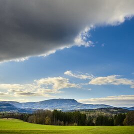 Weite Landschaft mit Blick auf den Schöckl, grüne Wiesen, Wälder und dramatische Wolkenstimmung.
