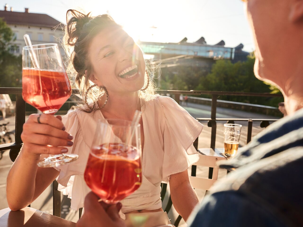 Menschen genießen Aperitifs auf einer Terrasse mit Blick auf das Kunsthaus Graz in der Abendsonne.