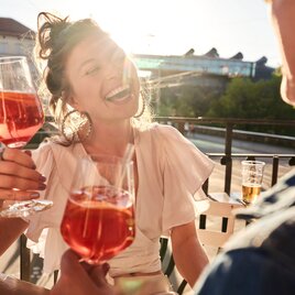 Menschen genießen Aperitifs auf einer Terrasse mit Blick auf das Kunsthaus Graz in der Abendsonne.