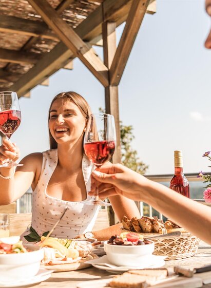 Drei Personen stoßen mit Schilcherwein an, sitzen an gedecktem Tisch mit Speisen auf sonniger Terrasse im Freien