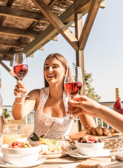 Three people toasting with Schilcher wine, sitting at a laid table with food on a sunny outdoor terrace