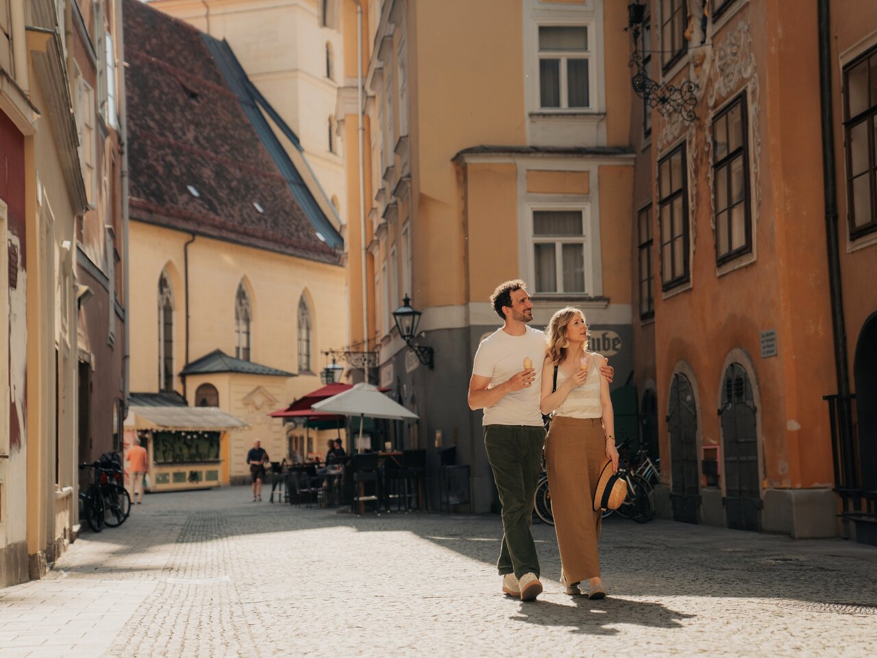 A young couple walks through a narrow historic old-town street, holding ice cream in warm sunlight.