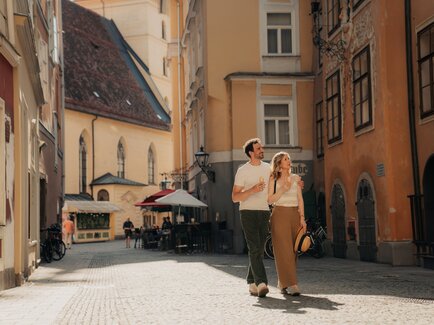 A young couple walks through a narrow historic old-town street, holding ice cream in warm sunlight.