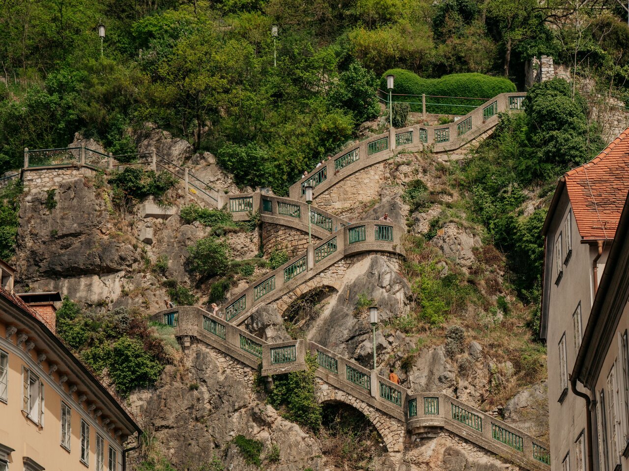 Blick auf die imposante Schlossbergtreppe in Graz, die sich in kunstvollen Windungen den steilen Fels hinaufzieht, flankiert von historischen Gebäuden und dem Eingang zur Veranstaltungsstätte „Dom im Berg“.