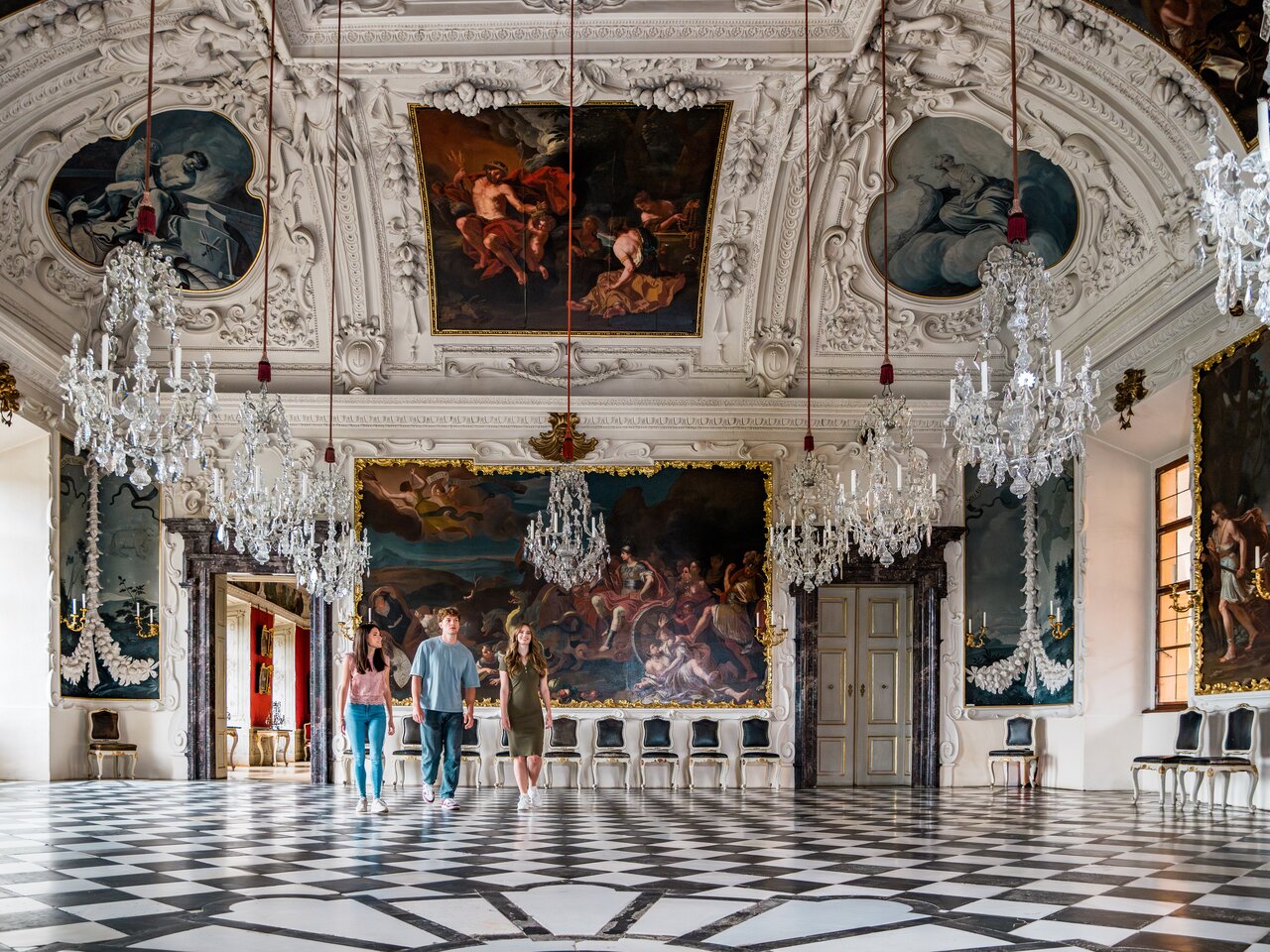 Barocker Prunkraum in Schloss Eggenberg in Graz mit aufwendig gestalteter Decke, historischen Gemälden, Kronleuchtern und Besuchern im Raum