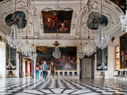 Barocker Prunkraum in Schloss Eggenberg in Graz mit aufwendig gestalteter Decke, historischen Gemälden, Kronleuchtern und Besuchern im Raum